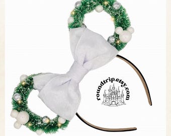 May include: White velvet bow headband with two green Christmas wreaths decorated with white and silver ornaments. The headband has the text "roundtrip.etsy.com" on it.