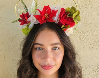 May include: A gold headband with a Mickey Mouse ear shape, decorated with red, white, and green flowers. The headband is worn by a person with long brown hair.