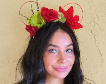 May include: A red and green floral headband with two circular wire loops. The headband is worn by a person with long dark hair.