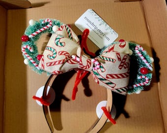 May include: A pair of Christmas-themed Minnie Mouse ears with a bow made of fabric with a candy cane pattern and green and white wreaths with red berries and white beads. The ears are attached to a brown headband.