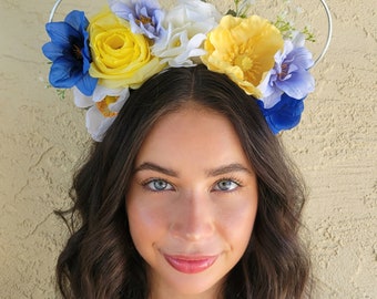 May include: A silver headband with blue and yellow flowers. The headband is decorated with blue, yellow, and white flowers. The flowers are arranged in a circle around the headband.