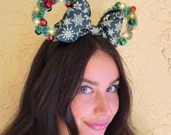 May include: A pair of green and red Christmas-themed Minnie Mouse ears with a black bow and white snowflake pattern. The ears are decorated with green tinsel, red berries, and small white lights.