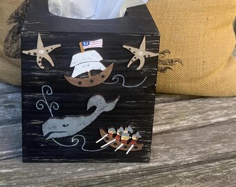 May include: A black wooden tissue box cover featuring nautical-themed cutouts including two beige starfish, a brown sailing ship with a small American flag, a grey whale, and a small boat with several figures rowing with white oars.