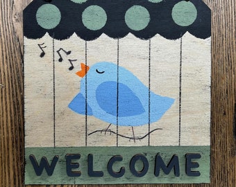 May include: A wooden welcome sign with a blue bird singing and green polka dots on a black background. The sign says "WELCOME" in black letters.