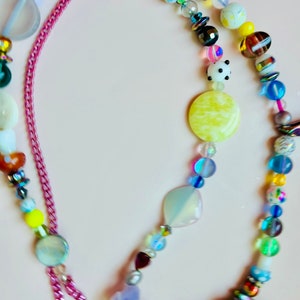 May include: A colorful beaded necklace with a pink chain clasp. The necklace features a variety of beads in different shapes, sizes, and colors, including white daisy-shaped beads, yellow, blue, and iridescent beads. The necklace is on a light pink background.