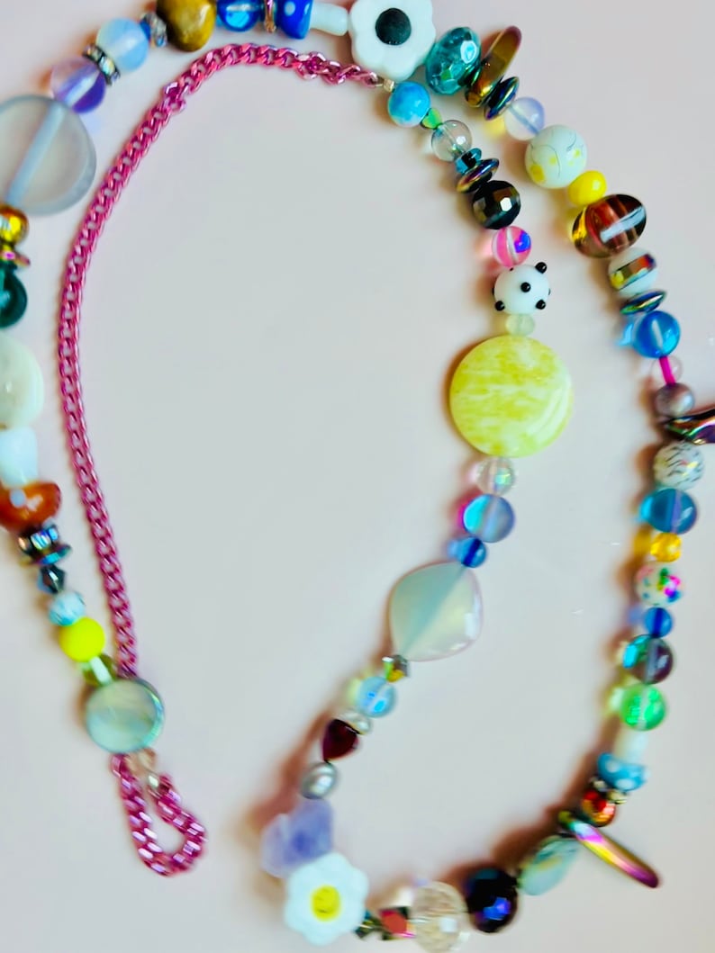 May include: A colorful beaded necklace with a pink chain clasp. The necklace features a variety of beads in different shapes, sizes, and colors, including white daisy-shaped beads, yellow, blue, and iridescent beads. The necklace is on a light pink background.