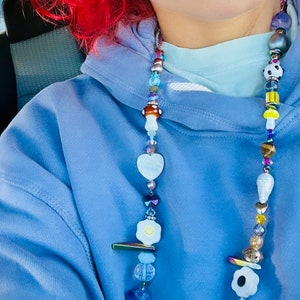 May include: A beaded necklace with a variety of colorful beads, including mushroom, heart, and flower shapes. The necklace features a mix of blue, white, yellow, and iridescent beads. The necklace is worn over a blue hoodie.