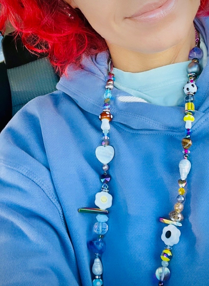 May include: A beaded necklace with a variety of colorful beads, including mushroom, heart, and flower shapes. The necklace features a mix of blue, white, yellow, and iridescent beads. The necklace is worn over a blue hoodie.