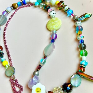 May include: A colorful beaded necklace with a variety of beads in different shapes, sizes, and colors. The necklace includes a chain, a flower with a smiley face, and a panda bear bead. The background is a light pink color.