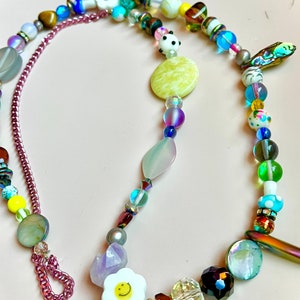 May include: A colorful beaded necklace with a variety of beads in different shapes, sizes, and colors. The necklace includes a chain, a flower with a smiley face, and a panda bear bead. The background is a light pink color.