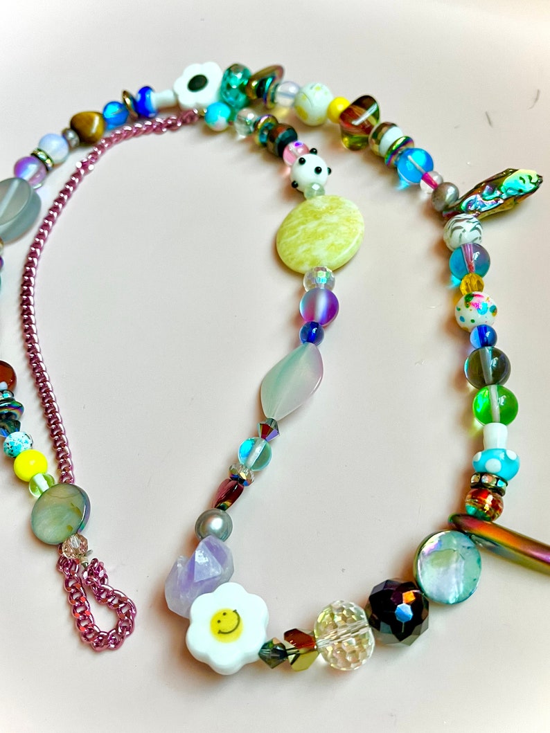 May include: A colorful beaded necklace with a variety of beads in different shapes, sizes, and colors. The necklace includes a chain, a flower with a smiley face, and a panda bear bead. The background is a light pink color.