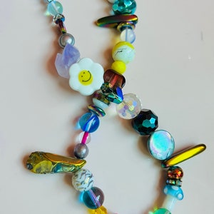 May include: A colorful beaded necklace featuring a variety of beads in different shapes, sizes, and colors. The necklace includes white daisy-shaped beads with yellow smiley faces, iridescent, and clear beads. The necklace is approximately 18 inches long.