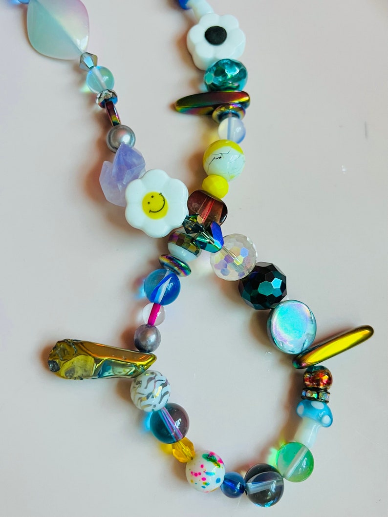 May include: A colorful beaded necklace featuring a variety of beads in different shapes, sizes, and colors. The necklace includes white daisy-shaped beads with yellow smiley faces, iridescent, and clear beads. The necklace is approximately 18 inches long.