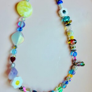 May include: A colorful beaded necklace featuring a variety of shapes and sizes. The necklace includes yellow, white, blue, and iridescent beads, with heart, flower, and geometric shapes. The necklace is displayed on a light pink surface.