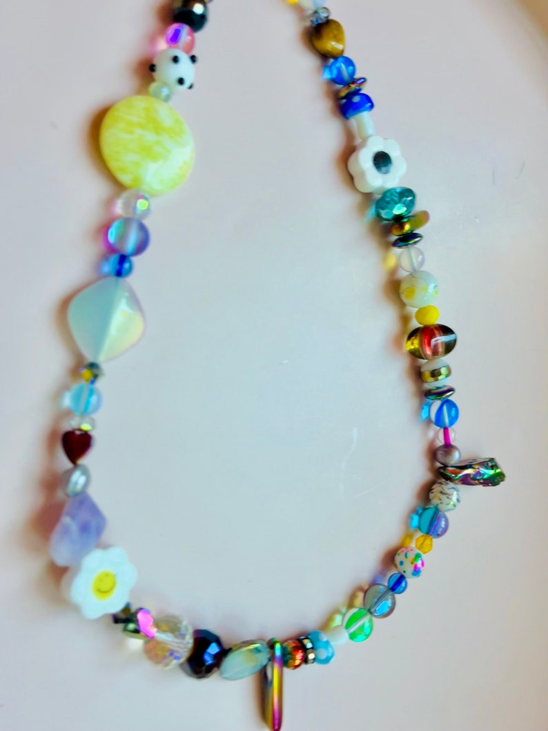 May include: A colorful beaded necklace featuring a variety of shapes and sizes. The necklace includes yellow, white, blue, and iridescent beads, with heart, flower, and geometric shapes. The necklace is displayed on a light pink surface.
