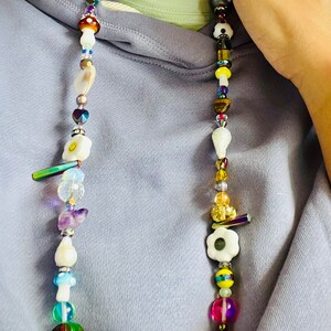 May include: A colorful beaded necklace with a variety of shapes and sizes. The necklace features mushroom, flower, and smiley face charms, along with iridescent and clear beads. The necklace is worn against a light purple hoodie.