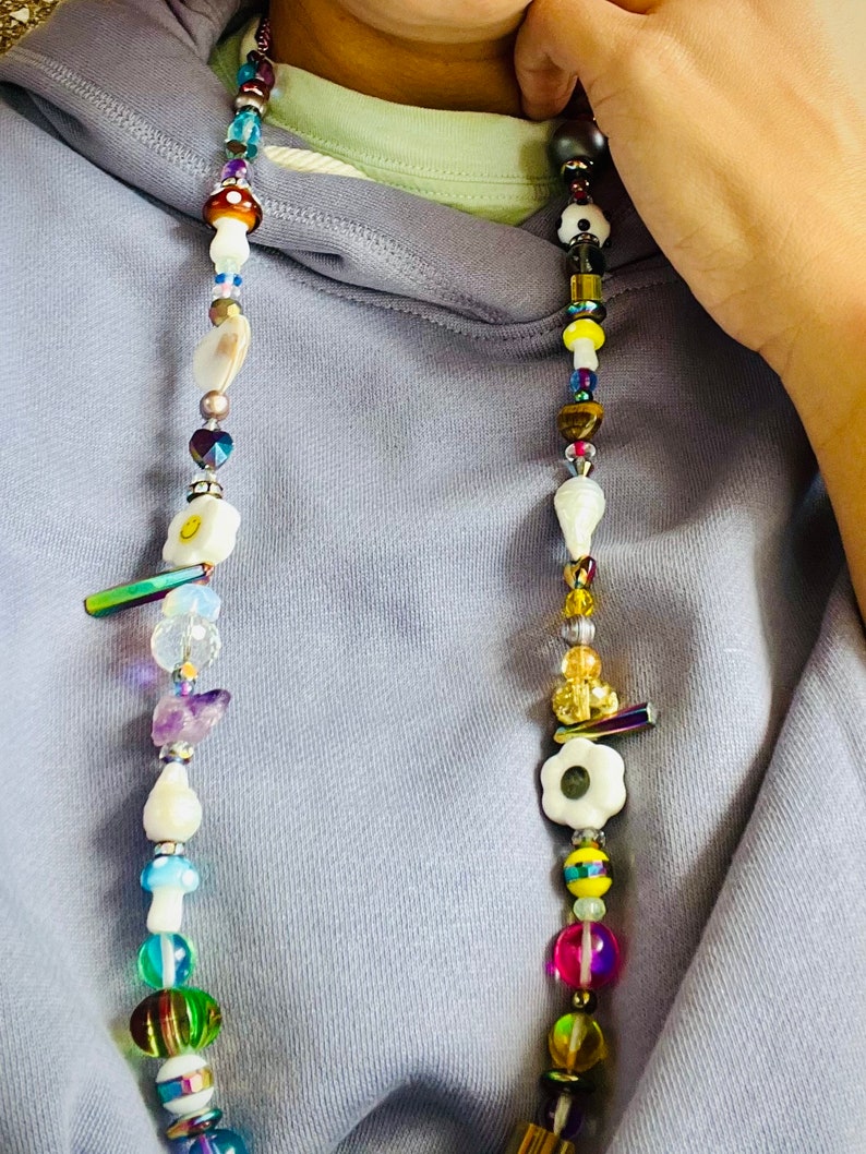 May include: A colorful beaded necklace with a variety of shapes and sizes. The necklace features mushroom, flower, and smiley face charms, along with iridescent and clear beads. The necklace is worn against a light purple hoodie.