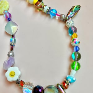 May include: A colorful beaded necklace with a variety of shapes and colors. The necklace features iridescent, clear, and opaque beads, including a white daisy with a smiley face, a yellow heart, and a mushroom-shaped bead. The necklace is on a pink background.