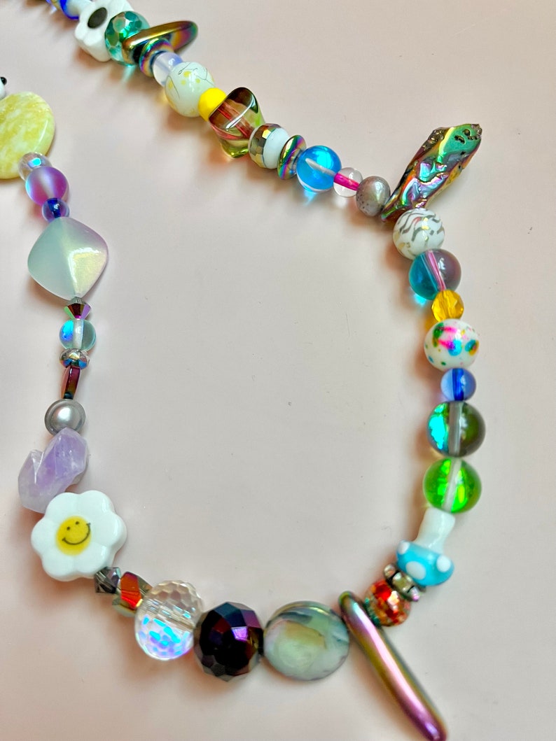 May include: A colorful beaded necklace with a variety of shapes and colors. The necklace features iridescent, clear, and opaque beads, including a white daisy with a smiley face, a yellow heart, and a mushroom-shaped bead. The necklace is on a pink background.