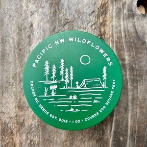 May include: A green round metal container with a white design of a forest scene with a tent, trees, and a sun. The text on the container reads "Pacific NW Wildflowers Decker Rd. Seeds Est. 2012 - 1 oz. Covers 250 Square Feet."