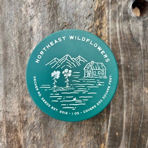 May include: A green round tin with white text and a line drawing of a house, mountains, and flowers. The text reads "Northeast Wildflowers Decker Rd. Seeds Est. 2012 - 1 oz. Covers 250 Square Feet."