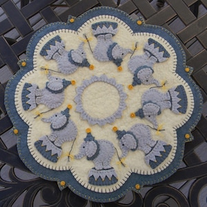 Può includere: Un arazzo da parete in feltro con un design circolare blu e bianco che presenta otto elefanti grigi con cappelli da festa e in equilibrio su palle. Gli elefanti tengono corde con stelle gialle all'estremità. Lo sfondo è di colore crema.