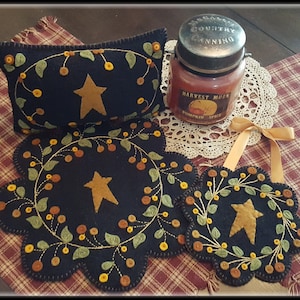 May include: Three black felt quilted ornaments with a star and berry design. The ornaments are on a plaid tablecloth with a candle and a doily.