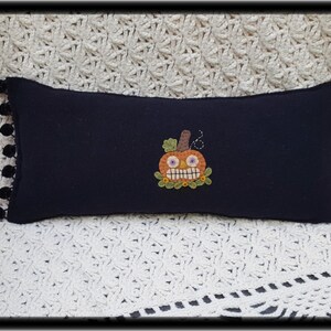 Puede incluir: Una almohada negra con ribete de pompones blancos presenta una calabaza naranja bordada con una sonrisa de dientes y ojos saltones.