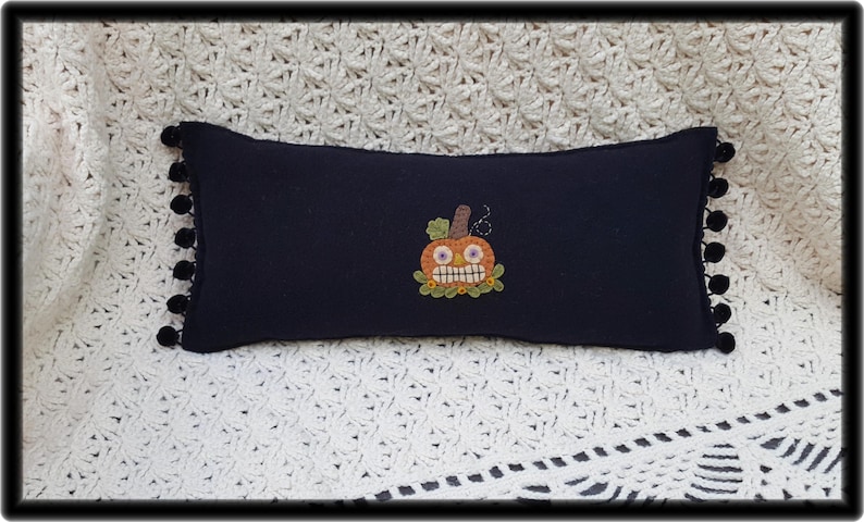 Puede incluir: Una almohada negra con ribete de pompones blancos presenta una calabaza naranja bordada con una sonrisa de dientes y ojos saltones.
