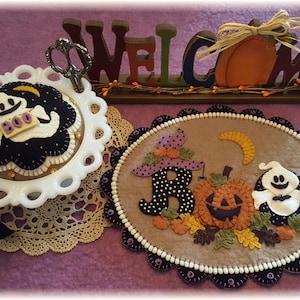 May include: A Halloween-themed felt craft with a ghost, pumpkin, and the word "BOO" in black and white. The craft is surrounded by a brown felt border with a white and black scalloped edge.