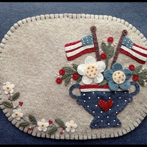 May include: A felt oval placemat with a patriotic theme. It features a blue polka-dotted vase with a red heart, holding flowers and two American flags. Additional floral accents and a stitched border complete the design.