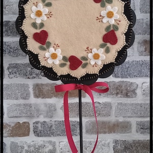 May include: A felt wall hanging with a scalloped edge and a cream-colored center. The center is decorated with embroidered white flowers and red hearts. The hanging is mounted on a black stand with a red ribbon.