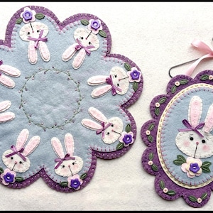 May include: Two felt bunny wall hangings with purple and blue accents. The larger one is a round, scalloped design with seven bunnies around the edge. The smaller one is oval shaped with a single bunny in the center.