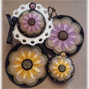 May include: Three felt pincushions with flower designs. The pincushions are in shades of purple, yellow, and brown. The pincushions are on a white background.