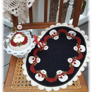 May include: A black felt snowman-themed table runner with white felt snowmen and red felt accents. The runner is on a wooden chair with a woven seat. A white felt pincushion with a snowman design is on the chair.