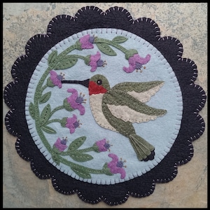 Puede incluir: Aplicación de fieltro de colibrí con fondo azul claro, rodeada por un borde negro festoneado. El colibrí es verde, gris y rojo, con un largo pico negro. También se presentan flores moradas y hojas verdes.