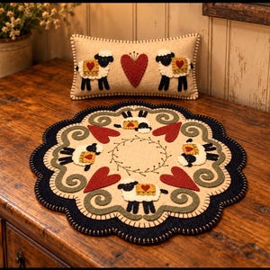 May include: A rectangular beige felt cushion featuring two black and white sheep with red heart blankets flanking a central red heart, and a round beige felt mat with a scalloped dark blue edge, decorated with four sheep, red hearts, and green swirls.