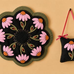 Puede incluir: Un tapete circular de fieltro con borde festoneado verde oliva y centro negro que presenta flores rosas similares a margaritas con centros naranjas y tallos verdes, junto a un pequeño adorno cuadrado de fieltro negro colgante.