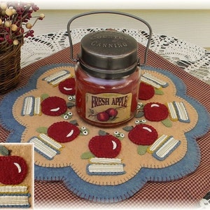 May include: A felt apple and books themed candle mat with a red apple and stack of books design. A glass jar candle with a metal lid and a "Fresh Apple" label is on top of the mat.