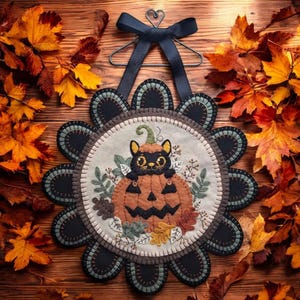 May include: A black cat peeking out of a felt pumpkin with a black and green floral design. The pumpkin is surrounded by fall leaves and is hanging from a black ribbon.