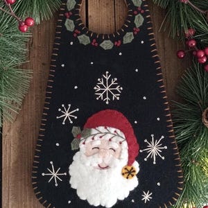 May include: A black felt door hanger featuring a hand-stitched Santa Claus face with a red hat, white beard, and a small yellow bell. The design includes embroidered white snowflakes, a holly leaf border around the top hole, and brown blanket stitching.