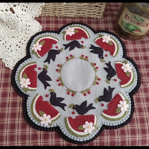 May include: A round, gray felt table runner with a black, white, and red watermelon design. The runner is decorated with black birds, white flowers, and red cherries.
