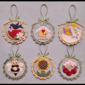 May include: Six felt ornaments with different designs, including a watermelon, a birdhouse, a kite, a sunflower, a bee, and cherries. Each ornament is attached to a green ribbon and has a scalloped edge.