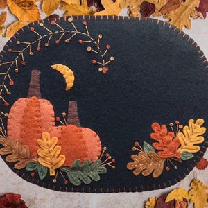 May include: Oval-shaped, dark blue felt placemat with embroidered autumn-themed design. Features orange pumpkins, a crescent moon, and fall leaves in shades of orange, yellow, and green. The edge is stitched with orange thread.