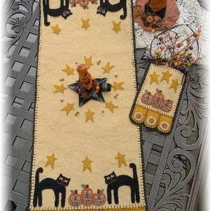May include: A beige table runner with a black cat, stars, and pumpkins. The runner has a scalloped edge with a brown and yellow pattern. A small matching piece with a similar design is also pictured.