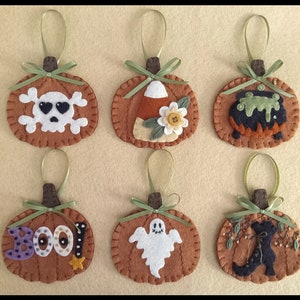 May include: Six felt pumpkin ornaments with different Halloween themes. The ornaments are brown with green ribbon and a brown stem. The themes include a skull and crossbones, a candy corn, a cauldron, the word "BOO!" with purple letters and glitter, a ghost, and a black cat with a witch's boot.