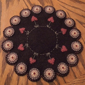 May include: A dark brown, circular felt trivet with a scalloped edge. The trivet features a ring of decorative, circular felt elements in shades of grey, pink, and brown. Small red felt hearts are evenly spaced around the centre circle, connected by embroidered stems.