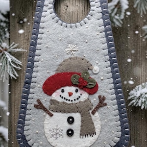 May include: A light gray felt door hanger featuring a snowman with a red hat, brown scarf, and two black buttons. The snowman is decorated with embroidered snowflakes and small white beads. The hanger has a dark gray border with white stitching.