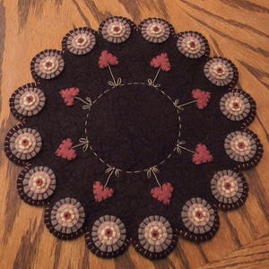 May include: A brown felt circle rug with a scalloped edge. The rug is decorated with pink and white felt flowers and pink felt hearts.