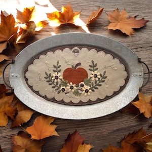 May include: A brown and beige felt pumpkin with white flowers and green leaves on a metal tray. The tray is oval shaped and has two handles. The tray is surrounded by fall leaves.